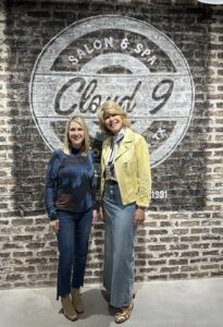 Stephanie Wilson and Kim Cloud inside Cloud 9 Salon and Spa in front of the Cloud 9 logo on a dark brick wall.